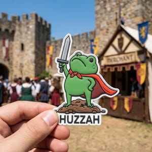 May include: A green frog sticker with a red cape and a silver sword, standing on a brown base with the word "Huzzah" below. The frog has a red bow tie and a stern expression. The background shows a medieval fair.