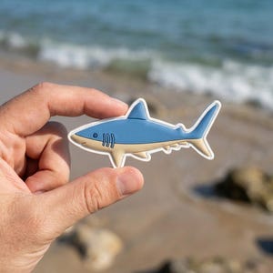 May include: A blue and tan shark-shaped sticker with a white outline. The sticker is held in a hand, with the ocean and beach in the background. The shark has a light blue body, tan belly, and white teeth.