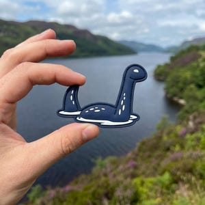 May include: A navy blue Nessie sticker with white accents, held in front of a lake and mountain backdrop. The sticker depicts the Loch Ness Monster with a long neck and a fin, floating in white water.