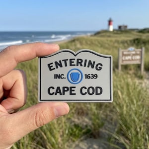 May include: A gray and black sign that reads "ENTERING CAPE COD" with the text "INC. 1639" and a blue emblem. The sign is held up in front of a beach scene with a lighthouse in the background.