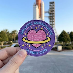 Puede incluir: Una pegatina redonda morada con el texto "GALACTIC AMBASSADOR" y "PLANET CUTE". La pegatina presenta un corazón rosa con un anillo amarillo y estrellas. El fondo incluye un transbordador espacial y árboles.