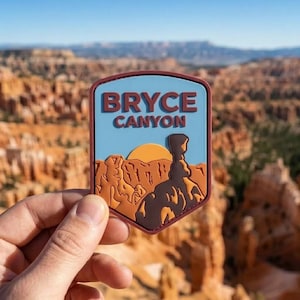 May include: A rectangular patch with the words "BRYCE CANYON" in white letters on a blue background. The patch features a landscape illustration with a setting sun and rock formations in shades of brown and orange.