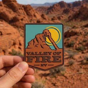 May include: A rectangular patch featuring the words "Valley of Fire - NV" in brown lettering. The patch has a teal background with a brown rock formation and a yellow sun. The patch is held in front of a blurred desert landscape.