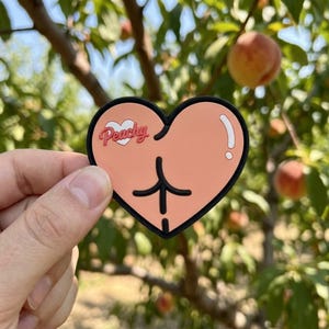 May include: A heart-shaped sticker with a peach-colored design and a black border. The sticker features a peach-colored buttock graphic and the word "Peachy" in a pink script. The background shows a peach tree with ripe peaches.