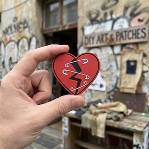 May include: A red heart-shaped patch with a black jagged line and three safety pins. The patch is held up in front of a weathered wall with graffiti and a sign that reads "DIY ART & PATCHES".