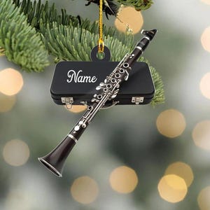May include: A clarinet ornament with a black case and the word "Name" in white script. The clarinet is dark brown with silver keys and is suspended by a gold string, perfect for a musician or music lover.