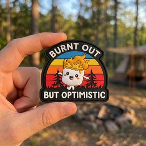 May include: A black patch with the words "BURNT OUT" and "BUT OPTIMISTIC" in white. The patch features a cartoon marshmallow character with flames on its head, set against a sunset background with trees.