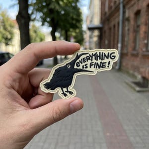 May include: A black crow-shaped patch with the text "EVERYTHING IS FINE!" in white. The crow has a long beak and is standing on a beige background. The patch is held in a hand.