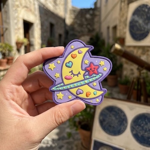 May include: A purple star-shaped sticker featuring a yellow crescent moon with a smiling face, surrounded by small stars and hearts. A red star is positioned near the moon. The sticker has a green ring around the moon and star.