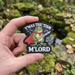 May include: A black patch featuring a cartoon frog wearing a crown, cape, and holding a sword. The text "T'WAS THE 'TISM" and "M'LORD" are displayed in white. The frog is green, the cape is red, and the sword is silver.