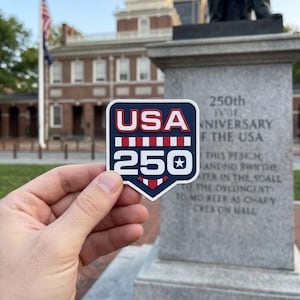May include: A patriotic sticker with a blue border and the text "USA 250" in red and white. The sticker commemorates the 250th anniversary of the USA. The background features a historic building and a monument.