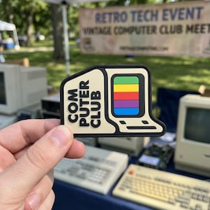 May include: A beige and black patch in the shape of a vintage computer. The computer screen displays a rainbow design. The words "COM PUTER CLUB" are printed on the left side of the patch. The background includes vintage computers and a banner that reads "Retro Tech Event".