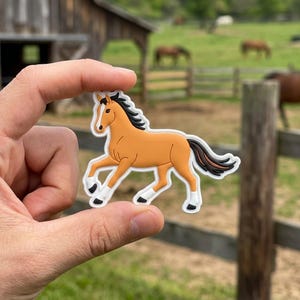 May include: A brown horse-shaped charm with a black mane and tail, white hooves, and a tan body. The charm is held between fingers against a blurred background of a barn and a pasture with horses.
