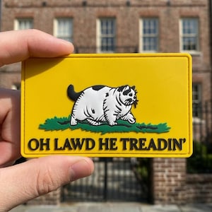May include: Yellow rectangular patch featuring a black and white cat walking on green grass. The text "OH LAWD HE TREADIN'" is printed in black below the cat illustration.