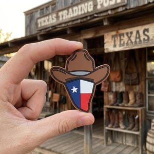 May include: A brown cowboy hat-shaped emblem with a Texas flag design. The emblem is held up in front of a rustic wooden building with the words "Texas Trading Post" and "Texas" visible.