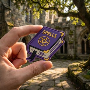 May include: A purple book-shaped charm with the word "SPELLS" in white lettering and a gold pentagram design. The charm has gold-colored edges and a black ribbon bookmark. The background is a blurred outdoor scene.