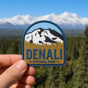 May include: A patch featuring the text "DENALI - NATIONAL PARK" in blue lettering. The patch has a blue arch with a mountain scene in white and black, set against a tan background. The patch is held up against a backdrop of mountains and trees.