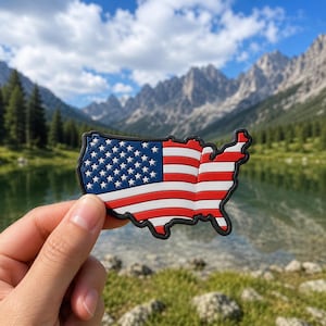 May include: A colorful magnet in the shape of the United States, featuring the American flag design. The magnet has a blue field with white stars and red and white stripes. The magnet is held in front of a scenic mountain and lake background.