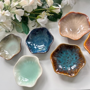 Pode incluir: Uma coleção de seis pequenas tigelas de cerâmica de formato irregular, em várias cores, incluindo azul, verde, castanho e laranja. As tigelas têm um acabamento brilhante e estão dispostas sobre uma superfície branca, com flores brancas ao fundo.