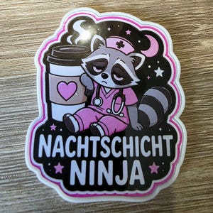 Könnte beinhalten: Ein gestanzter Aufkleber mit einem müden Waschbären in rosa medizinischer Kleidung und Krankenschwesterhaube, der einen Kaffeebecher mit Herzsymbol hält. Das Design zeigt ein Stethoskop, einen Halbmond, Sterne und den Text NACHTSCHICHT NINJA.
