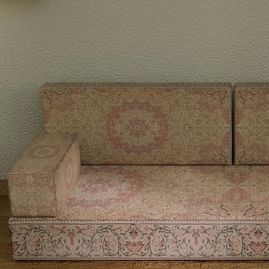 May include: A low-profile, patterned sofa with a beige and pink floral design. The sofa has two back cushions and two armrests. The base of the sofa features a decorative border. The sofa is placed on a wooden floor.