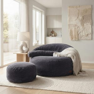 May include: A dark gray corduroy circular lounge chair and a matching round ottoman. The chair features a low profile, a wide seat, and a long cylindrical bolster pillow. A soft, cream-colored textured throw blanket is draped over the side.