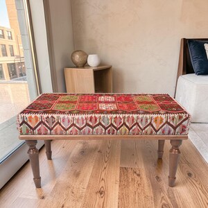 Puede incluir: Un otomán rectangular con una parte superior textil estampada y colorida. El textil presenta diseños geométricos rojos, verdes y marrones. El marco y las patas de madera son de color marrón cálido. El otomán está sobre un suelo de madera.