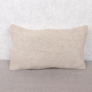 Puede incluir: Un cojín rectangular beige con un diseño tejido texturizado. El cojín está colocado sobre una superficie gris, contra un fondo gris claro. La forma del cojín es ligeramente curvada, lo que aumenta su atractivo visual.