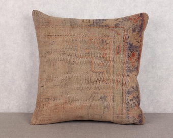 May include: Square cushion with a distressed, vintage appearance. The beige fabric is patterned with faded red, blue, and orange hues. The cushion, likely woven, adds a touch of rustic charm. Measurements are approximately 40cm x 40cm.