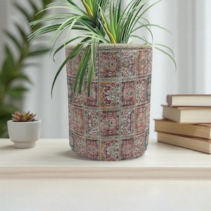 May include: A cylindrical plant pot with a detailed, patterned design in shades of red, beige, and green. The pot contains a green plant with long, slender leaves. The pot sits on a light-colored wooden surface.