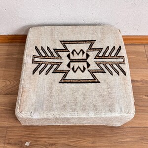 May include: Square beige ottoman with a geometric design in brown and black. The pattern is centered on the top surface. The ottoman is placed on a wooden floor.