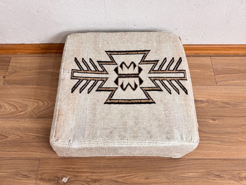 May include: Square beige ottoman with a geometric design in brown and black. The pattern is centered on the top surface. The ottoman is placed on a wooden floor.