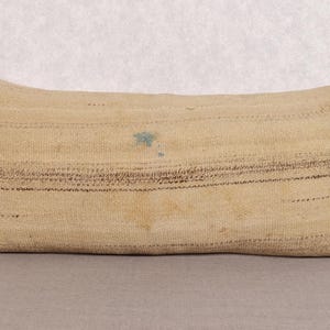 Puede incluir: Cojín rectangular beige con rayas horizontales marrones y una pequeña mancha azul. El cojín tiene una textura tejida y un detalle anudado en un extremo. Adecuado para una cama o sofá.