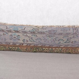 May include: Rectangular decorative pillow with a vintage rug design. The pillow features a faded color palette of blue, purple, and beige, with intricate floral and geometric patterns. The edges are trimmed with a brown and green border, adding a touch of rustic charm.