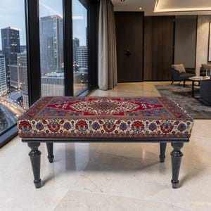 May include: A rectangular ottoman with a detailed, patterned top in red, cream, blue, and purple. The ottoman has a black wooden frame and four turned legs. The design is reminiscent of a traditional rug.