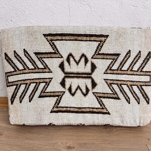 May include: Rectangular beige pillow with a geometric pattern in brown and tan. The design features symmetrical shapes and lines, creating a textured appearance. The pillow is placed on a wooden surface.