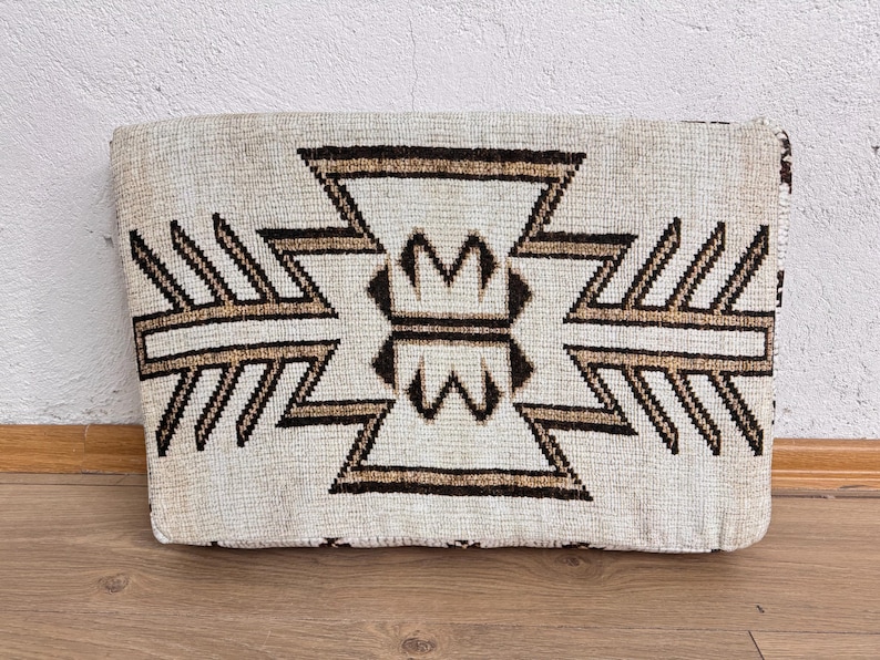 May include: Rectangular beige pillow with a geometric pattern in brown and tan. The design features symmetrical shapes and lines, creating a textured appearance. The pillow is placed on a wooden surface.
