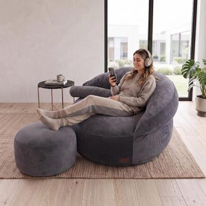 May include: A dark gray corduroy bean bag chair with a matching round ottoman. The chair features a rounded, deep-seated design with a soft, textured fabric finish. The set provides a low-profile seating arrangement with a cohesive, monochromatic appearance.