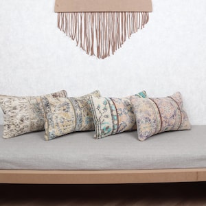 May include: Four rectangular throw pillows with vintage-style patterns in beige, blue, and red tones are arranged on a light gray upholstered bench. The pillows have a distressed look, suggesting an antique or bohemian aesthetic.