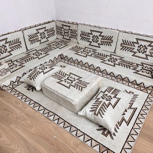 May include: An off-white sectional sofa with a geometric brown pattern. The sofa includes multiple cushions and a matching rug, all featuring the same design. The set is arranged on a wooden floor.