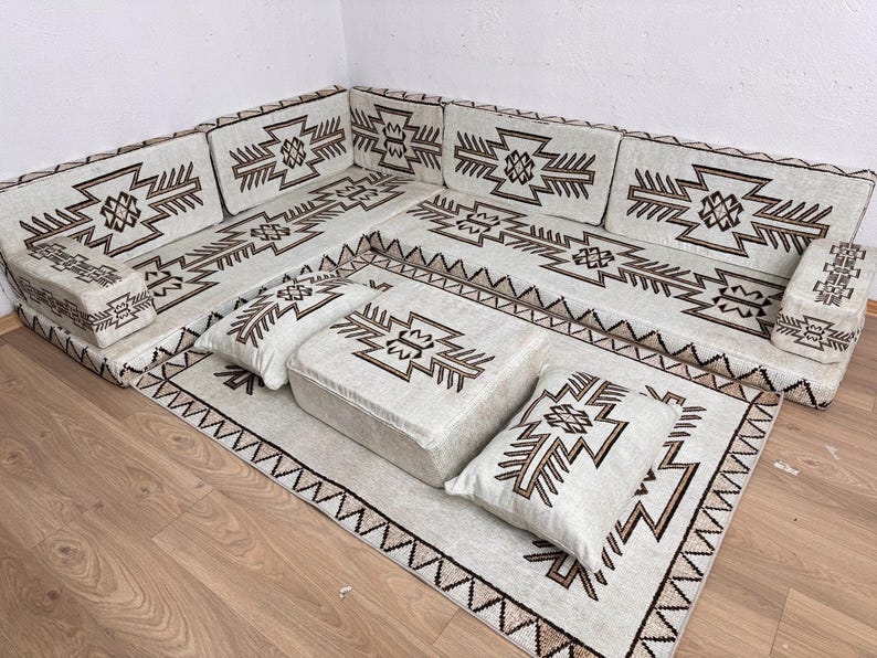 May include: An off-white sectional sofa with a geometric brown pattern. The sofa includes multiple cushions and a matching rug, all featuring the same design. The set is arranged on a wooden floor.