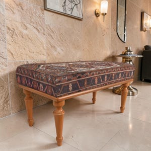 Op de afbeelding: Een rechthoekige bank met een patroontextiel bovenkant en lichtbruine houten poten. Het textiel heeft een complex ontwerp in tinten rood, bruin en blauw. De bank is een decoratief meubelstuk.