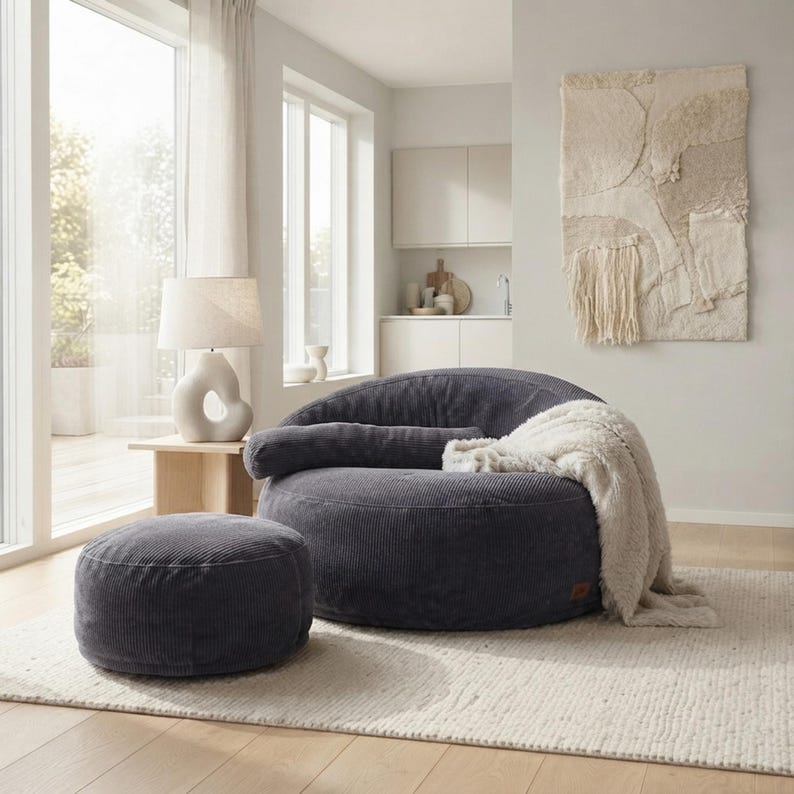 May include: A dark gray corduroy circular lounge chair and a matching round ottoman. The chair features a low profile, a wide seat, and a long cylindrical bolster pillow. A soft, cream-colored textured throw blanket is draped over the side.