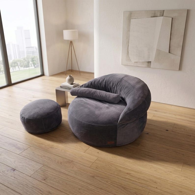 May include: A dark gray corduroy lounge chair with a rounded, low-profile silhouette and a matching cylindrical bolster pillow, accompanied by a small, circular, matching dark gray corduroy ottoman, both featuring a soft, textured fabric finish.