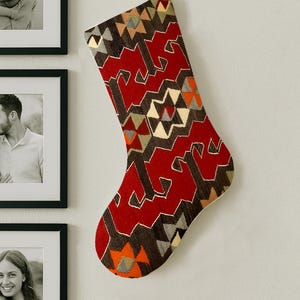 Puede incluir: Un calcetín navideño con un patrón geométrico en rojo, marrón, naranja y beige cuelga de una pared blanca. El calcetín tiene un diseño en zigzag y está hecho de tela tejida. En el fondo hay fotografías enmarcadas.