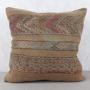 May include: Square decorative pillow with a woven design in shades of tan, brown, and pink. The pillow features geometric patterns and horizontal stripes, adding texture and visual interest to the design.