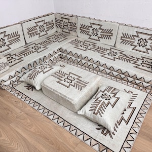 Puede incluir: Un juego de sofá seccional crema con una alfombra y otomana a juego. El conjunto presenta patrones geométricos marrones. La alfombra y las almohadas tienen diseños similares. El sofá está dispuesto en forma de L.
