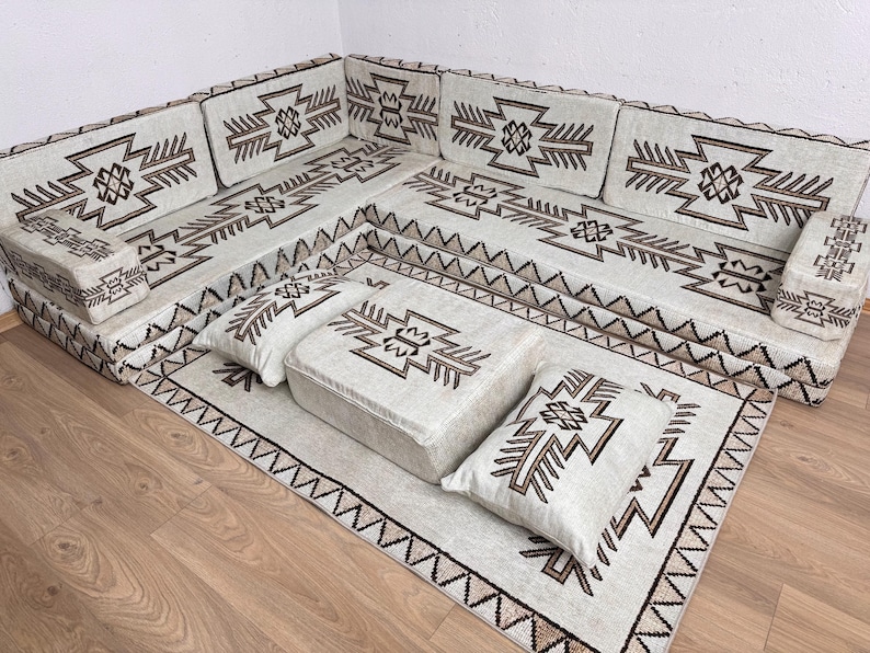 May include: A sectional sofa set with a matching rug and ottoman. The set features a cream-colored fabric with geometric brown patterns. The rug and pillows have similar designs. The sofa is arranged in an L-shape.