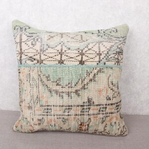 May include: Square decorative pillow with a vintage rug design. The pillow features a patchwork of muted colors, including beige, light blue, and touches of orange. The top section has a geometric pattern, while the bottom has a more intricate, faded design.