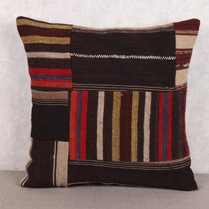 May include: Square decorative pillow with a patchwork design. The pillow features a mix of dark brown, beige, red, and blue vertical and horizontal stripes. The pillow is made of a woven fabric.